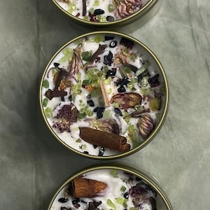 May include: Three round candles with a white wax base and various natural elements embedded in the wax, including dried flowers, cinnamon sticks, and green and black crystals. The candles are in metal containers with a gold-coloured finish.