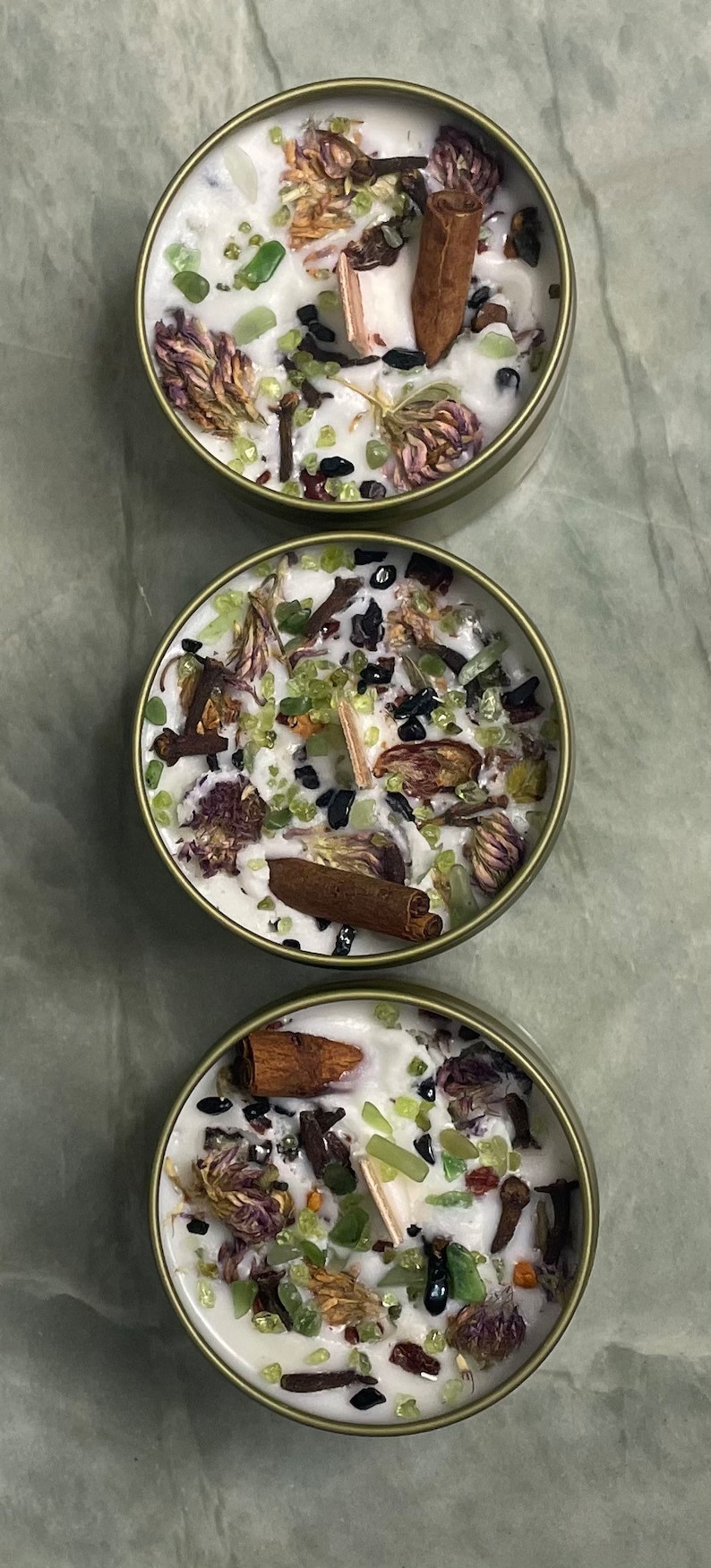 May include: Three round candles with a white wax base and various natural elements embedded in the wax, including dried flowers, cinnamon sticks, and green and black crystals. The candles are in metal containers with a gold-coloured finish.
