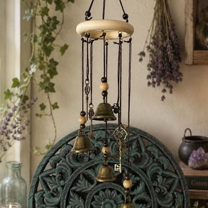 Op de afbeelding: Een hangende windgong met messing bellen, houten kralen en decoratieve sleutels. De windgong is opgehangen aan een houten ring en zwarte koorden. De achtergrond omvat een gesneden houten paneel en gedroogde lavendel.