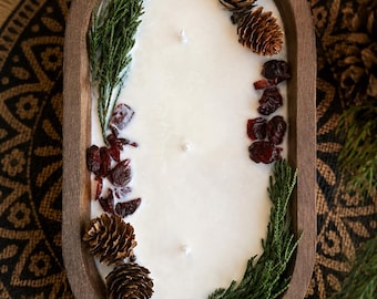 May include: An oval-shaped wooden dough bowl candle featuring white wax with three wicks. The surface is decorated with natural elements including small brown pinecones, dried red cranberries, and sprigs of green cedar foliage arranged along the edges.