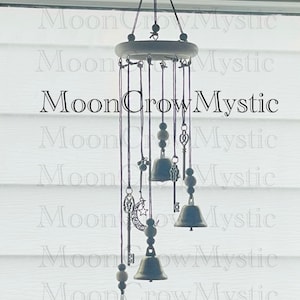 Op de afbeelding: Decoratieve windgong met een houten bovenkant en hangende metalen bellen. De windgong heeft bedels, waaronder sleutels, een maan en sterren. De tekst "MoonCrowMystic" is zichtbaar.