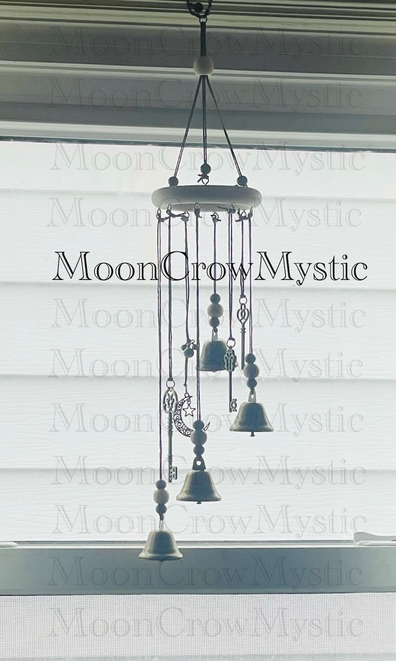 Op de afbeelding: Decoratieve windgong met een houten bovenkant en hangende metalen bellen. De windgong heeft bedels, waaronder sleutels, een maan en sterren. De tekst "MoonCrowMystic" is zichtbaar.