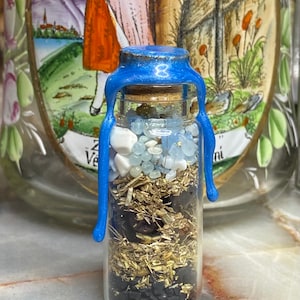May include: A small glass bottle with a blue wax seal and cork stopper. The bottle is filled with various crystals, herbs, and other materials. The bottle is sitting on a light brown surface.