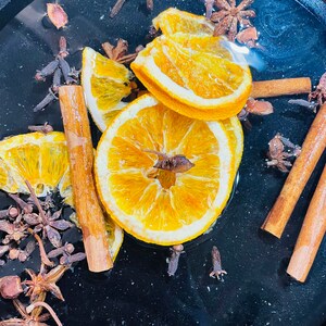 Puede incluir: Un cuenco negro lleno de agua y rodajas de naranja, palitos de canela y clavos. Las rodajas de naranja est&aacute;n secas y tienen un color amarillo. Los palitos de canela son marrones y los clavos son marr&oacute;n oscuro.