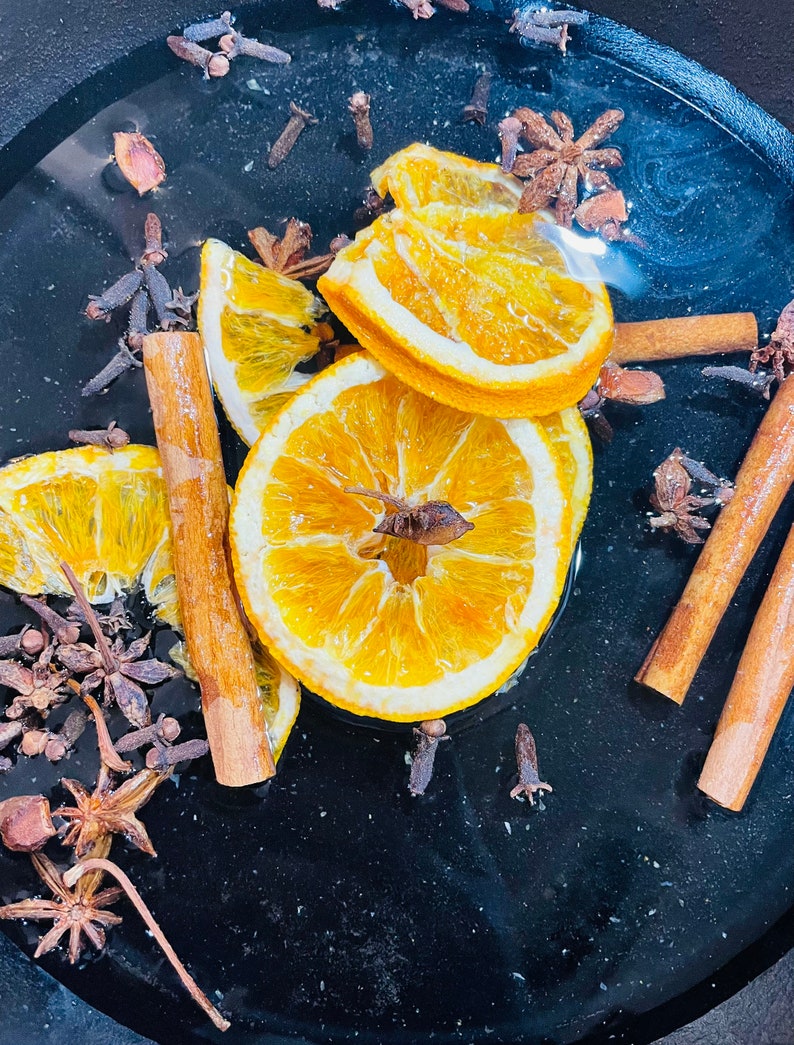 Puede incluir: Un cuenco negro lleno de agua y rodajas de naranja, palitos de canela y clavos. Las rodajas de naranja est&aacute;n secas y tienen un color amarillo. Los palitos de canela son marrones y los clavos son marr&oacute;n oscuro.