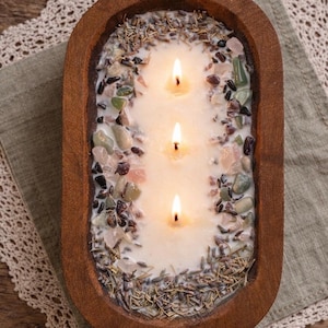 May include: A three-wick candle, alight, in an oval wooden bowl. The candle is surrounded by small stones and dried herbs. The wooden bowl sits on a neutral-coloured surface. The candle is a decorative item.