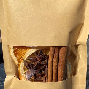 Puede incluir: Una bolsa de papel marr&oacute;n con una ventana transparente que muestra rodajas de naranja secas, an&iacute;s estrellado y palitos de canela en su interior.