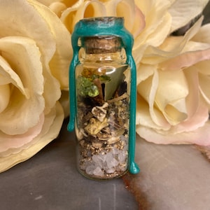 Op de afbeelding: Een kleine glazen fles gevuld met kruiden, kristallen en natuurlijke elementen. De fles is afgesloten met een kurk en teal wax. De achtergrond toont een zacht, wazig bloemstuk.