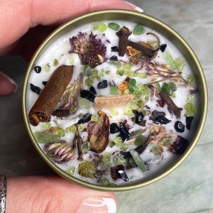 May include: A white candle with green, brown, and black herbs and crystals embedded in the wax. The candle is in a gold tin.