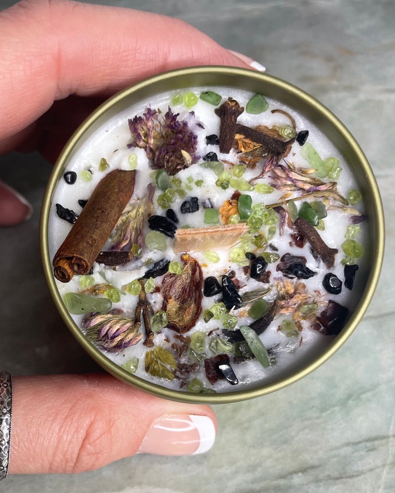 May include: A white candle with green, brown, and black herbs and crystals embedded in the wax. The candle is in a gold tin.