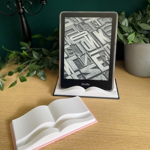 May include: A Kindle e-reader, showing a page of letterforms, sits on a white book-shaped stand. A second, smaller book-shaped stand with a pink border is in front. The background features a green wall and a candle holder.