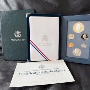May include: A 1990 United States Mint Prestige Set including a dark green outer box, a white display folder with red, white, and blue ribbon accents, a blue coin display case containing six coins, and a Certificate of Authenticity document.