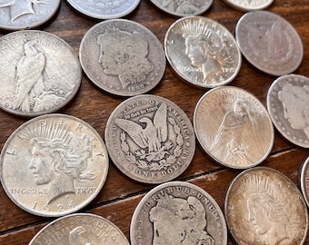 May include: A collection of silver US coins featuring various designs, including an eagle, a woman's profile, and the words "In God We Trust" and "One Dollar".