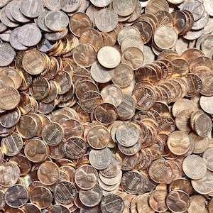 Op de afbeelding: Een close-up afbeelding van een grote stapel koperkleurige Amerikaanse centen. De munten zijn in een willekeurig patroon gerangschikt, waarbij sommige munten elkaar overlappen. De afbeelding is gericht op de munten, en de achtergrond is wazig.