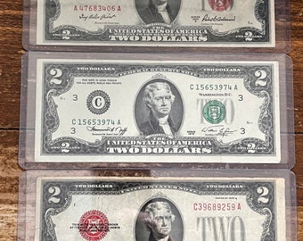 Könnte beinhalten: Drei US-Zwei-Dollar-Scheine in Schutzhüllen. Jeder Schein zeigt ein Porträt von Thomas Jefferson und die Worte „TWO DOLLARS“. Die Scheine stammen aus verschiedenen Serienjahren mit unterschiedlichen Seriennummern und Siegeln. Die Scheine werden auf einer Holzoberfläche präsentiert.
