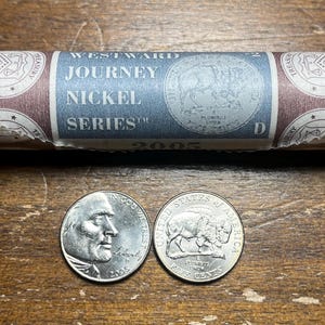 May include: A roll of 2005 Westward Journey Nickel Series coins. The roll is wrapped in brown paper with the words "Westward Journey Nickel Series" and "2005" printed on it. The roll is sitting on a wooden surface. Two coins are sitting in front of the roll. One coin features a portrait of a man, and the other coin features a buffalo.