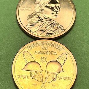 May include: Two golden-colored United States one-dollar coins. The top coin features Sacagawea with her child and the text LIBERTY and IN GOD WE TRUST. The bottom coin displays two military helmets, feathers, and the text UNITED STATES OF AMERICA, $1, WWI, WWII, and CODE TALKERS.