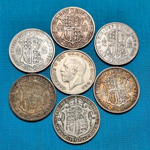 May include: A collection of antique silver coins, including half crowns and a coin featuring a profile of a man. The coins display intricate designs, including heraldic emblems and text. The coins are arranged on a blue surface.