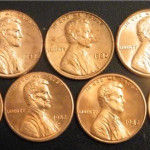 May include: Seven copper-colored Lincoln pennies, each displaying a profile of Abraham Lincoln. The coins are dated 1982, with the word "LIBERTY" and the phrase "IN GOD WE TRUST" visible. The pennies are arranged on a dark surface.