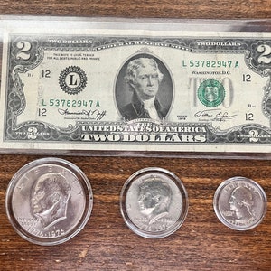 Può includere: Una banconota da due dollari del 1976 con numero di serie L53782947 A. La banconota è in una custodia di plastica. Nell'immagine ci sono tre monete d'argento, due sono in custodie di plastica e una no. Le monete sono tutte a fuoco.