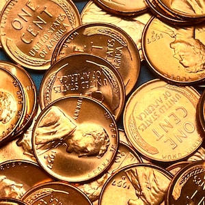 May include: Close-up of a pile of United States one-cent coins. The copper-colored pennies display the words "One Cent", "United States of America", and the year "1957". The image shows the profile of Abraham Lincoln.