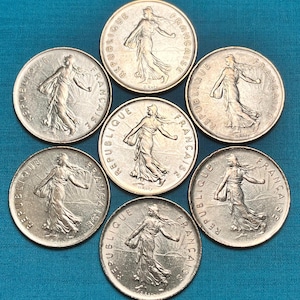 May include: Seven silver French coins with the inscription "REPUBLIQUE FRANCAISE" and the image of a woman walking. The coins are arranged in a circle on a blue surface.