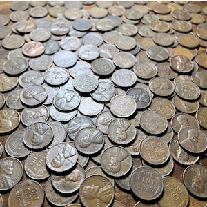 May include: A large collection of United States one-cent coins, also known as pennies, are scattered across a surface. The copper-colored coins display the profile of Abraham Lincoln and the words "One Cent" and "United States of America."