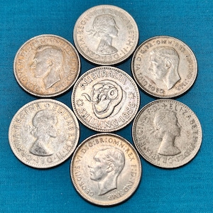 May include: A collection of vintage silver coins, including Australian shillings and coins featuring portraits of Queen Elizabeth II and King George VI. The coins are arranged on a blue surface.