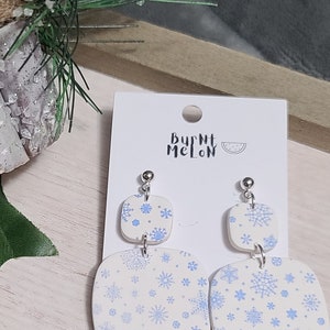 May include: A pair of white snowflake earrings with blue snowflake designs. The earrings are on a white card with the text "Burnt Melon" and a watermelon graphic.