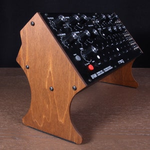 May include: A black synthesizer with numerous knobs and buttons is mounted on a wooden stand. The stand is made of light brown wood and has a curved design. The synthesizer has the brand name "Moog" on it. The stand is secured with screws.