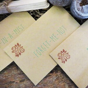 Op de afbeelding: Drie rechthoekige zaadzakjes van beige papier met groene tekst en een kleine bruine bloemenstempel. De zakjes zijn gelabeld met Love-in-a-Mist, Forget-Me-Not en Poppy. Elk zakje toont een consistent botanisch ontwerp in bruine inkt.