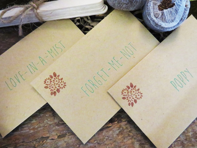 Op de afbeelding: Drie rechthoekige zaadzakjes van beige papier met groene tekst en een kleine bruine bloemenstempel. De zakjes zijn gelabeld met Love-in-a-Mist, Forget-Me-Not en Poppy. Elk zakje toont een consistent botanisch ontwerp in bruine inkt.