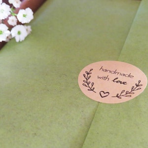 Op de afbeelding: Een ovale, beige sticker met de tekst "handmade with love" in zwarte inkt, voorzien van een decoratief bladontwerp aan beide zijden en een klein hartje onderaan, geplakt op een gestructureerd olijfgroen papieren oppervlak.