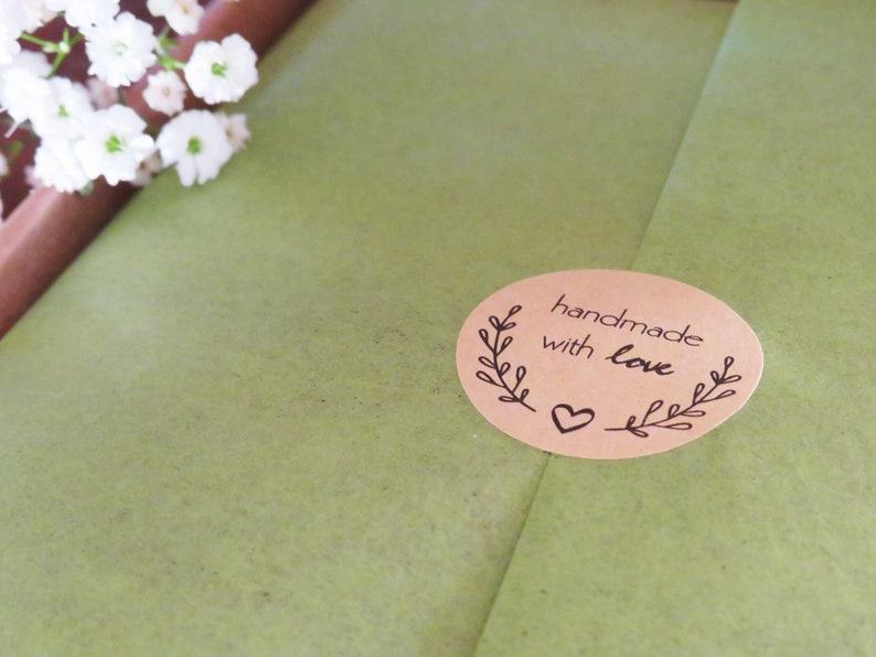 Op de afbeelding: Een ovale, beige sticker met de tekst "handmade with love" in zwarte inkt, voorzien van een decoratief bladontwerp aan beide zijden en een klein hartje onderaan, geplakt op een gestructureerd olijfgroen papieren oppervlak.