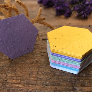 May include: A stack of hexagonal seed paper in various pastel colours, including yellow, green, blue, and purple. The paper is made from recycled materials and is designed to be planted.