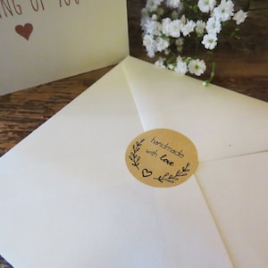 Op de afbeelding: Een cr&egrave;mekleurige envelop verzegeld met een ronde bruine sticker met de tekst handmade with love, een klein hartje en bladillustraties. Op de achtergrond toont een gedeeltelijke wenskaart de tekst THINKING OF YOU met een hartje.