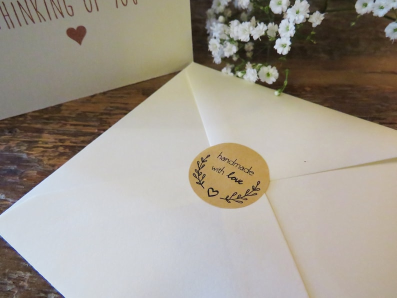 Op de afbeelding: Een cr&egrave;mekleurige envelop verzegeld met een ronde bruine sticker met de tekst handmade with love, een klein hartje en bladillustraties. Op de achtergrond toont een gedeeltelijke wenskaart de tekst THINKING OF YOU met een hartje.