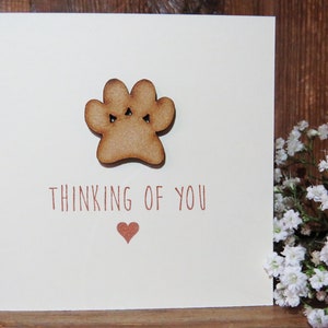 Op de afbeelding: Een vierkante wenskaart met een cr&egrave;mekleurige achtergrond en een houten pootafdruk-uitsnede in het midden bovenaan. Onder de pootafdruk staat de tekst "THINKING OF YOU" in bruin gedrukt, gevolgd door een klein bruin hartsymbool onder de tekst.