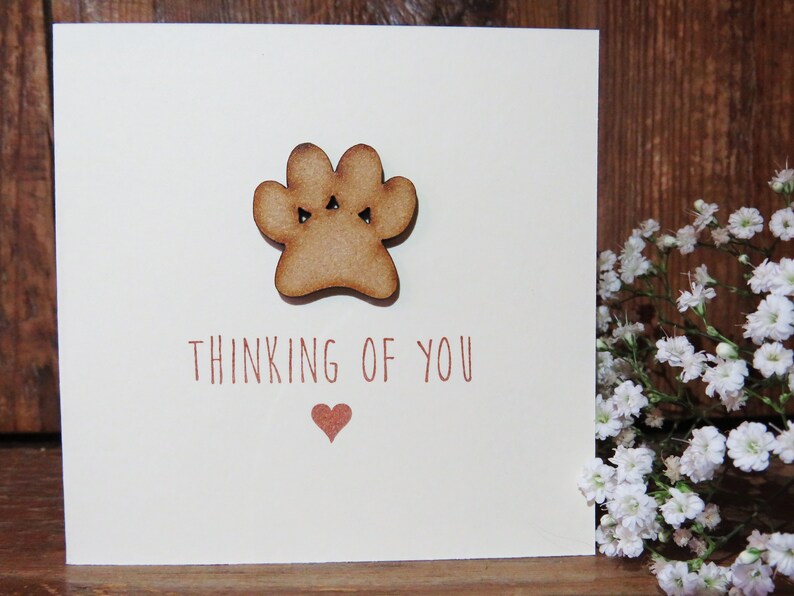 Op de afbeelding: Een vierkante wenskaart met een cr&egrave;mekleurige achtergrond en een houten pootafdruk-uitsnede in het midden bovenaan. Onder de pootafdruk staat de tekst "THINKING OF YOU" in bruin gedrukt, gevolgd door een klein bruin hartsymbool onder de tekst.