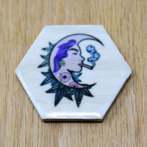 May include: A hexagonal wooden coaster with a colorful illustration of a woman with purple hair smoking a cigarette in a crescent moon shape surrounded by green leaves.