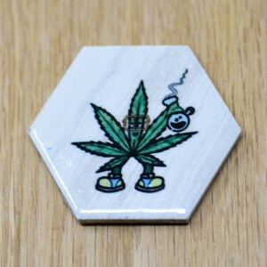 May include: A white hexagonal coaster with a green marijuana leaf design and a cartoon character smoking a pipe. The character is wearing blue and yellow shoes.