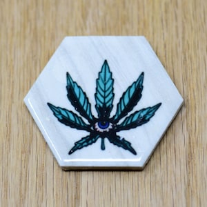 May include: A white hexagonal coaster with a blue and black marijuana leaf design with an eye in the center.