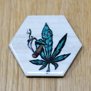 May include: A hexagonal ceramic coaster with a white background and a blue marijuana leaf design. The leaf has a cartoon face and is holding a joint.