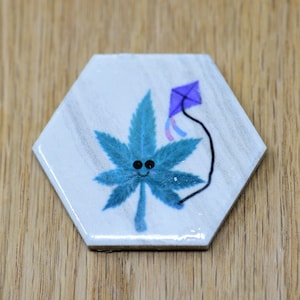 May include: A hexagonal ceramic coaster with a white background and a blue and green marijuana leaf with a smiling face holding a purple kite.