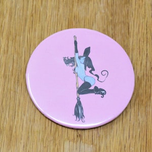 May include: A pink round button with a black and white illustration of a witch riding a broomstick. The witch is wearing a blue outfit and a black hat.
