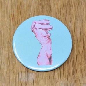 Könnte beinhalten: Ein runder, hellblauer Knopf mit einer rosa Illustration einer stehenden Person, die die Arme über den Kopf erhoben hat.