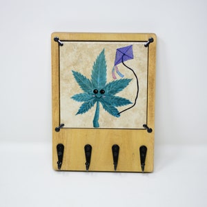 May include: A wooden key holder with a tile featuring a blue and green marijuana leaf with a smiling face and a purple kite. The key holder has four black metal hooks.