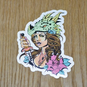 May include: A sticker featuring a colorful illustration of a woman with long brown hair wearing a green dragon head as a hat. She is holding a dagger in her right hand and a pink rose is behind her.