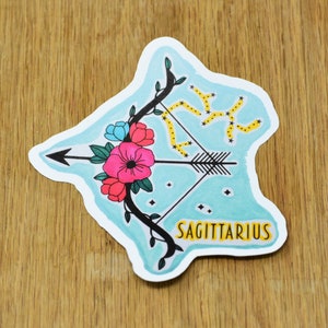 May include: A blue and white sticker with a black and white illustration of a bow and arrow with flowers. The constellation Sagittarius is depicted in yellow and the word "Sagittarius" is written in yellow letters.