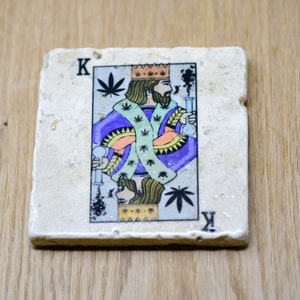 May include: A stone coaster with a printed image of a king of hearts playing card. The king is wearing a purple robe with a crown and holding a glass pipe. The card has a green marijuana leaf design.